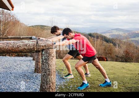 Vue latérale des joggings mâles fatigués bleus et rouges chemises qui s'étirent sur une clôture en bois après une course et un entraînement difficiles sur une colline verdoyante avec un magnifique paysage de montagne en arrière-plan Banque D'Images