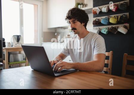 Vue latérale indépendante masculine, assise à une table dans la cuisine et travailler sur le projet en utilisant l'ordinateur Banque D'Images