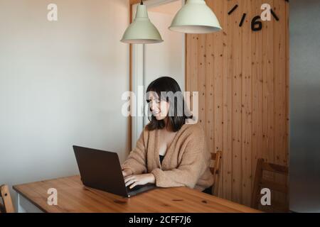 Femme pigiste souriante dans un confortable chandail assis à table cuisine et travail sur le projet tout en utilisant l'ordinateur Banque D'Images