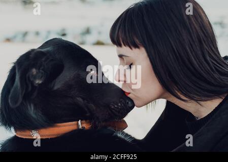 Vue latérale de la jeune femme brunette avec les yeux fermés embrassant dans le nez adorable chien noir Banque D'Images