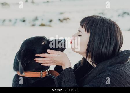 Vue latérale de la jeune femme brunette avec les yeux fermés embrassant dans le nez adorable chien noir Banque D'Images