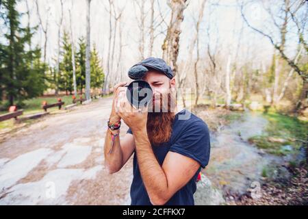 Homme moderne à tête rouge barbu en casquette prenant des photos avec l'appareil photo sur la route entourée d'arbres sans feuilles dans des couleurs vives bois le jour ensoleillé de l'automne Banque D'Images