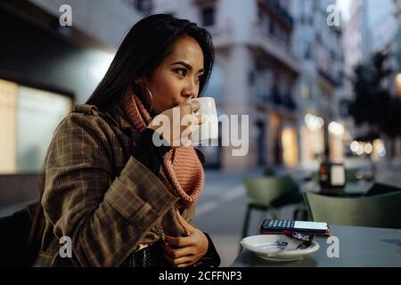 Femme ethnique dans l'extérieur tendance sirotant une boisson chaude fraîche et regardant loin tout en s'asseyant à table et en se reposant dans le restaurant de rue en soirée Banque D'Images