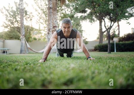 D'un niveau inférieur à un solide homme mature et actif regardant la caméra à l'intérieur bonne forme sportive planant et poussant vers le haut dans le pré vert dans le parc en été Banque D'Images