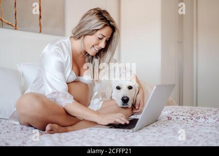 Femme enceinte blonde souriante avec chemise et soutien-gorge blancs ouverts taper sur un ordinateur portable tout en étant assis à la maison sur le lit avec jambes croisées et tête de chien du labrador debout à côté et regarder l'appareil photo Banque D'Images