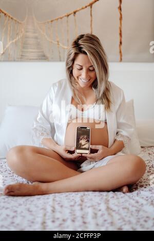 Femme enceinte montrant l'image du bébé hans à l'aide d'une échographie sur un smartphone, assis dans un soutien-gorge et une chemise ouverte sur un lit aux jambes croisées Banque D'Images