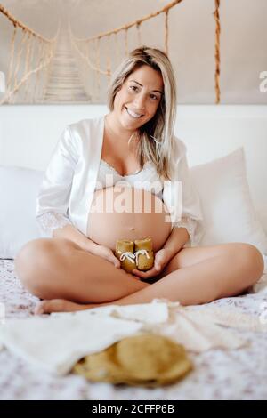 Femme enceinte vêtue d'un chemisier blanc ouvert et d'un soutien-gorge montrant des vêtements pour enfant à naître assis sur un lit aux jambes croisées Banque D'Images