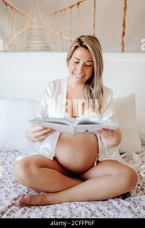 Contenu blonde femme enceinte vêtue blanc élégant maison vêtements de lecture réservez en vous asseyant avec des jambes croisées sur le lit dans des couleurs vives chambre Banque D'Images