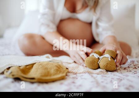 Femme enceinte sans visage vêtue de blouse blanche ouverte et soutien-gorge avec vêtements pour enfants à naître assis sur le lit jambes croisées Banque D'Images