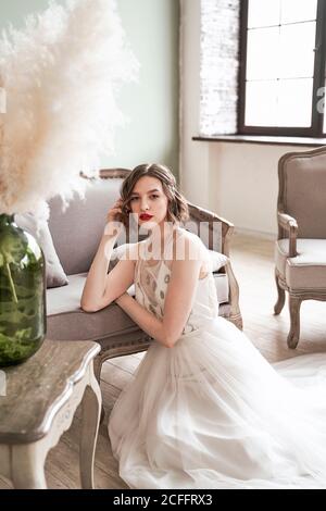 Magnifique femme avec des lèvres rouges dans la robe blanche regardant l'appareil-photo tout en étant assis sur le sol à côté du canapé Banque D'Images