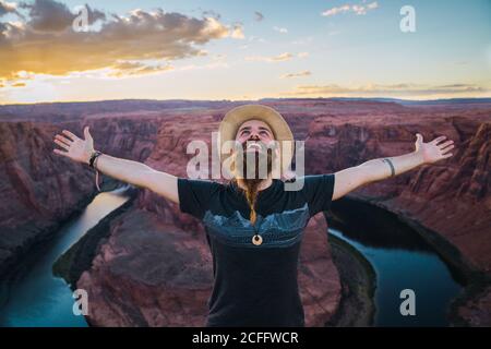 Beau homme en chapeau étirant les bras en regardant le ciel sur le majestueux canyon contre ciel bleu ciel nuageux pendant le voyage Par la côte ouest des États-Unis Banque D'Images