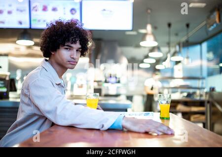 Vue latérale d'un jeune homme ethnique avec des cheveux bouclés à la caméra et en attendant la date tout en étant assis à table avec des verres sur le jus dans la cafétéria Banque D'Images
