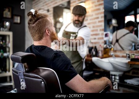 Coiffeur tailler la barbe d'un homme à tête rouge assis dans un salon de coiffure Banque D'Images