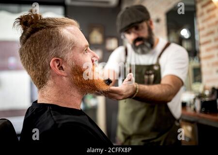 Coiffeur avec tondeuse barbe de redhead homme assis salon de coiffure Banque D'Images