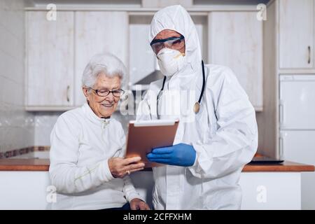 Médecin masculin méconnaissable en uniforme et masque de protection avec stéthoscope montrant et expliquant la prescription médicale à une femme âgée lors d'une visite chez le patient pendant l'éclosion du coronavirus Banque D'Images
