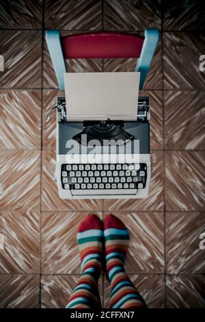Vue de dessus de l'auteur sans visage dans des chaussettes dénudées se tenant près chaise en bois sorbette avec machine à écrire à l'ancienne Banque D'Images