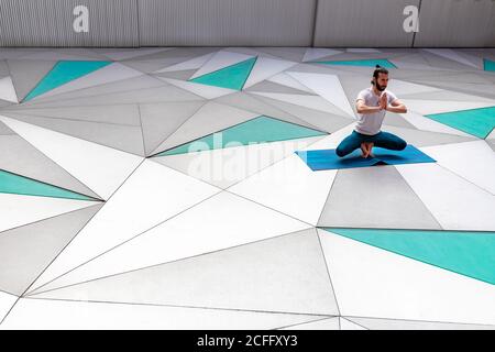 Un homme barbu en vêtements de sport s'entraînant sur l'équilibre des orteils avec geste de la mudra de prière dans une chambre spacieuse avec mur géométrique et au sol Banque D'Images