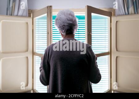Femme aux cheveux gris debout devant la fenêtre à la maison Banque D'Images