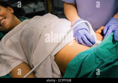 Une femme non reconnaissable a été coupée dans un gant bleu à l'aide d'un échographe, tout en examinant une femme enceinte gaie et en regardant un écran d'ordinateur à l'hôpital Banque D'Images