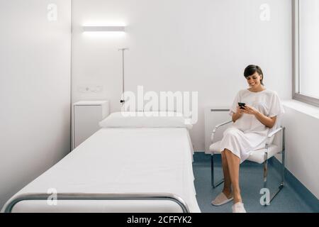 Femme gaie en robe blanche assise sur le lit près du lit et de la navigation de smartphone dans la salle de clinique moderne Banque D'Images