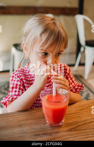 Appétissant verre parfumé de smoothie rouge dans les mains d'adorable enfant le buvant à table Banque D'Images