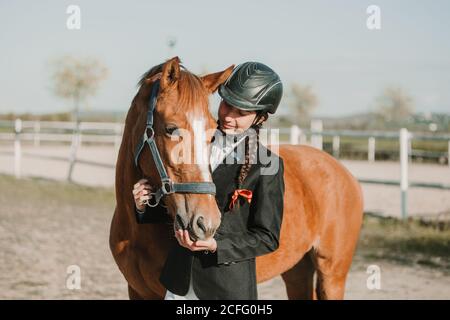 Vue latérale d'une jeune femme dans un casque de jockey et une veste caressant un cheval debout ensemble à l'extérieur Banque D'Images