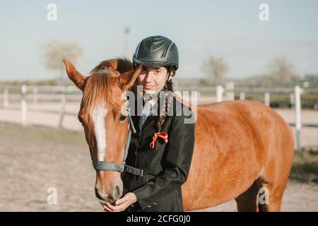 Vue latérale d'une jeune femme dans un casque de jockey et une veste caressant un cheval debout ensemble à l'extérieur et regardant la caméra Banque D'Images