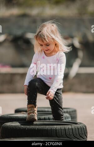 Bonne joyeuse petite fille ayant du plaisir et marchant dans la rangée de pneus de voiture noirs en jouant à l'extérieur le jour de l'été Banque D'Images