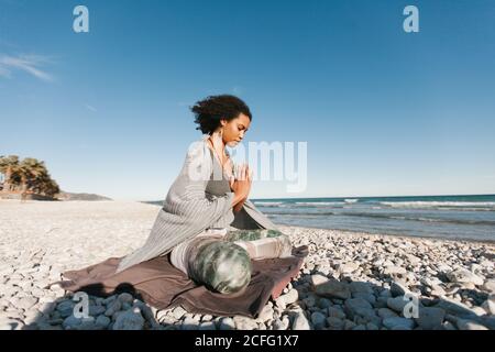 Vue latérale d'Afro-américaine jeune femme attrayante couverte de plaid gris méditant et priant dans lotus yoga posture sur la plage de sable en plein jour Banque D'Images