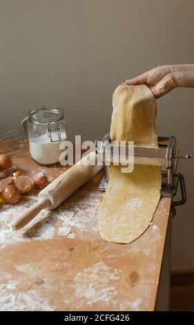 Femme sans visage roulant de pâtisserie avec machine à pâtes tout en cuisinant un délicieux dîner avec de la farine et des ustensiles de cuisine sur la table en bois à la maison Banque D'Images