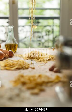 raw différents types de pâtes se trouvant sur une table en bois désordonné couvert de farine avec une bouteille d'huile d'olive et de coquilles d'œufs ustensiles de cuisine Banque D'Images