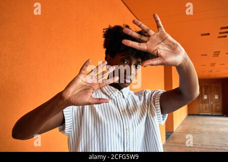 Femme afro-américaine adulte avec les yeux fermés essayant d'arrêter impact négatif en se tenant debout avec les bras relevés contre flou vif intérieur orange dans le couloir du bâtiment moderne Banque D'Images