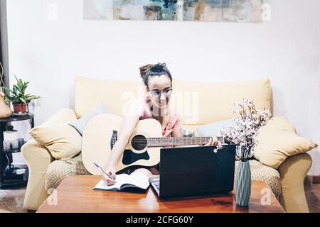 Magnifique femme prenant des notes tout en apprenant à jouer de la guitare à la maison avec des leçons en ligne sur son ordinateur portable. Banque D'Images