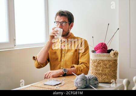 Homme adulte prenant un thé chaud frais et regardant loin pendant assis à la table près des aiguilles et des fils à tricoter Banque D'Images