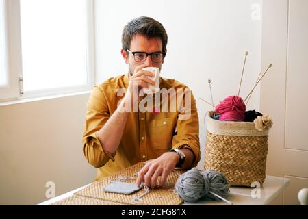 Homme adulte prenant un thé chaud frais et regardant loin pendant assis à la table près des aiguilles et des fils à tricoter Banque D'Images