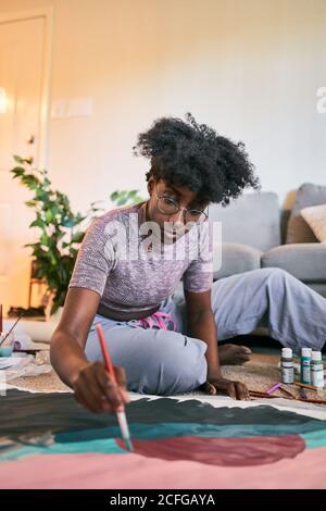 Jeune femme afro-américaine avec cheveux afro, artiste amateur dans une tenue décontractée dessin d'image colorée avec pinceau sur papier tout en étant assis sur le sol dans un appartement confortable Banque D'Images