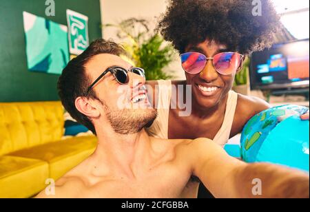 Joyeux couple multiracial portant des lunettes de soleil assis dans la piscine gonflable et prendre un selfie tout en profitant d'un week-end Banque D'Images