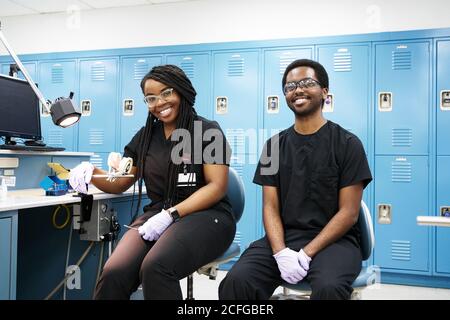 Bonne femme afro-américaine avec des tresses et un homme à barbe noire souriant et regardant la caméra tout en créant des prothèses dentaires dans un laboratoire moderne Banque D'Images