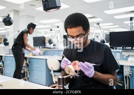 Afro-américain gars en latex gants utilisant le miroir de bouche et sonde permettant de vérifier les fausses dents lors de travaux dans un laboratoire moderne Banque D'Images