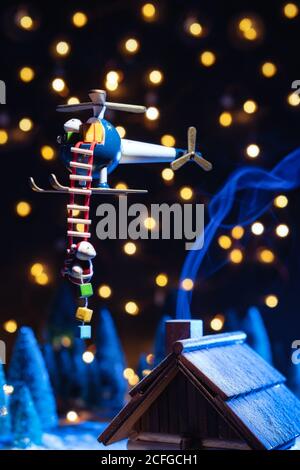 Petit Père Noël descendant de l'échelle de l'hélicoptère jouet au-dessus de la maison teintée la nuit d'hiver avec des lumières sur flou arrière-plan Banque D'Images