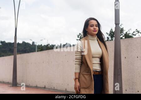 Jeune femme attirante à cheveux longs en col roulé léger et veste sans manches marchant dans la rue par mauvais temps Banque D'Images