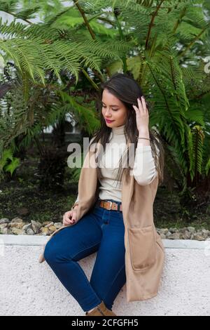 Jeune femme attirante à cheveux longs en col roulé léger et veste sans manches, assise sur une clôture en pierre et cheveux lissants Banque D'Images