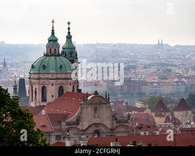 Prague depuis le château : une vue d'ensemble brumeuse tôt le matin de la ville et de ses clochers depuis le parc du château. Le dôme de Saint-Nicolas domine le premier plan et les doubles flèches noires de la basilique Saint-Pierre et de la basilique Saint-Paul sont à l'horizon. Banque D'Images