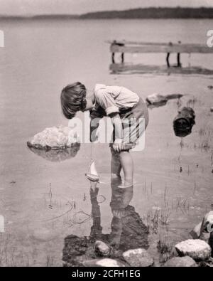 UN PETIT GARÇON DES ANNÉES 1920 JOUANT AVEC LE JOUET VOILE BATEAU PANT JAMBES ROULÉ SE PENCHER SUR DANS POND PAR LA RIVE VOIT SON RÉFLEXION DANS L'EAU - J289 HAR001 HARS SERENITY B&W FREEDOM SHORE FLEXION RÊVES BONHEUR GRAND ANGLE AVENTURE DÉCOUVERTE SES LOISIRS LES JEUNES ENFANTS DE LA CULTURE DE LA PATAUGEOIRE VOIENT LE NOIR ET L'ETHNIE BLANCHE CAUCASIENNE HAR001 ANCIEN MODE Banque D'Images