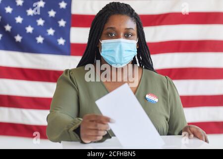 Portrait d'une jeune femme afro-américaine portant un masque mettant le bulletin de vote dans les urnes et regardant la caméra tout en se tenant contre le drapeau américain le jour de l'élection, espace de copie Banque D'Images