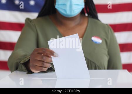 Gros plan d'une femme afro-américaine portant un masque mettant le bulletin de vote dans les urnes et regardant la caméra tout en se tenant contre le drapeau américain le jour de l'élection, espace de copie Banque D'Images