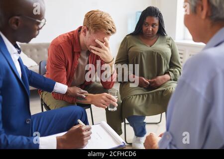 Portrait du jeune homme pleurant partageant ses problèmes pendant le soutien réunion de groupe avec des personnes qui le trouvent en cercle et le réconfortant Banque D'Images