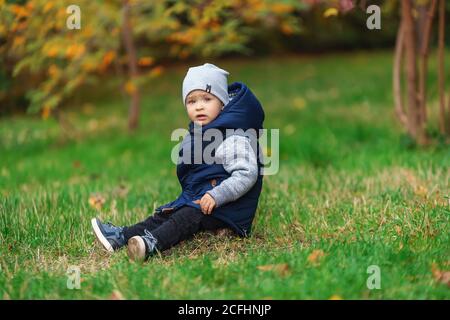 Portrait extérieur de bébé garçon à la mode dans le parc d'automne. Enfant dans une veste bleue et dans un chapeau gris assis sur l'herbe verte Banque D'Images
