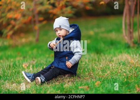 Un petit garçon à la mode jouant dans le parc d'automne. Enfant assis sur l'herbe verte Banque D'Images