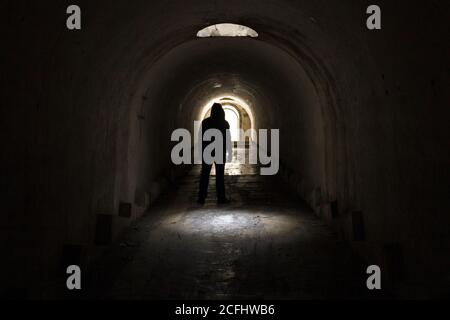 Un homme en vêtements sombres avec un capot allant à la sortie de la lumière d'un tunnel souterrain sombre, éclairé par la lumière solaire d'une trappe dans le plafond Banque D'Images